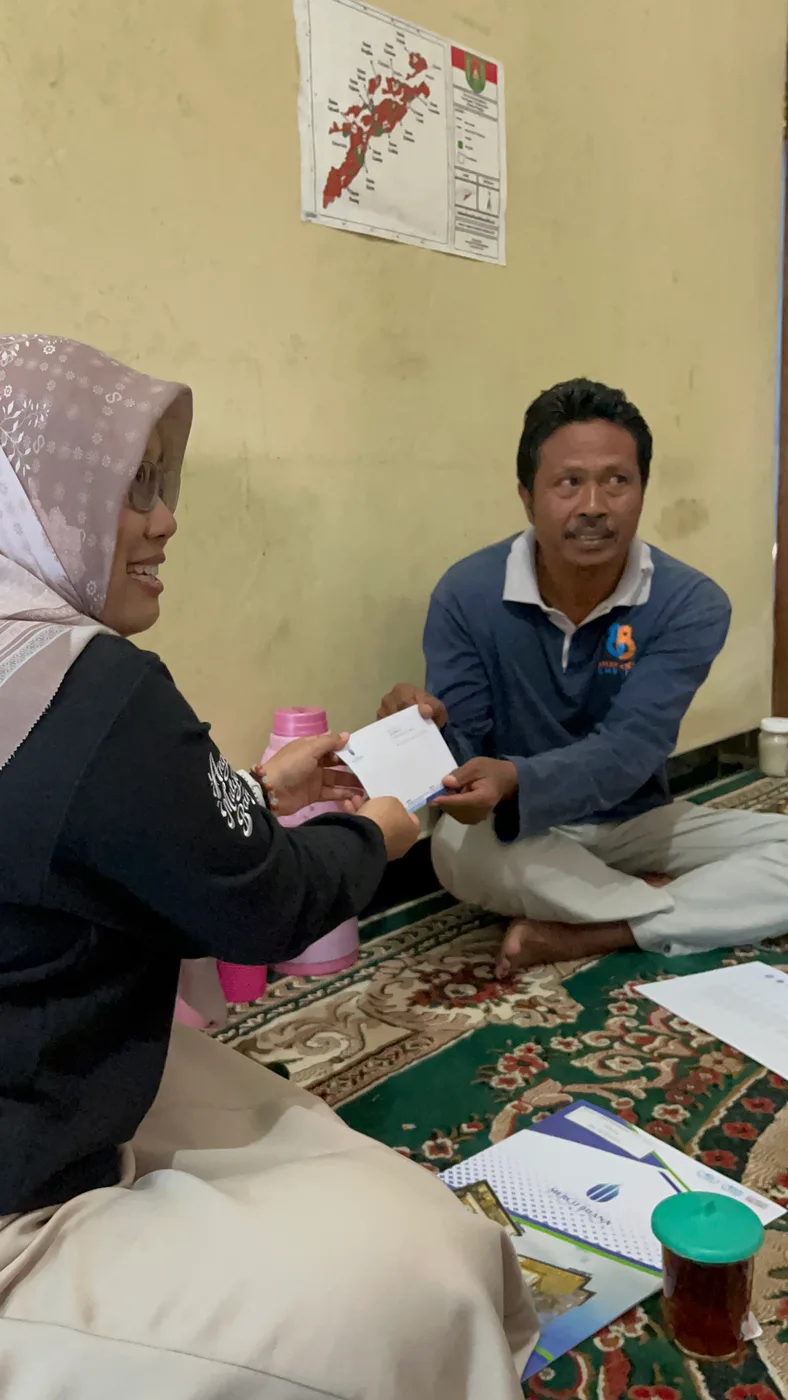 Seorang mahasiswa dan warga duduk bersila berhadapan sambil menyerahkan selembar dokumen