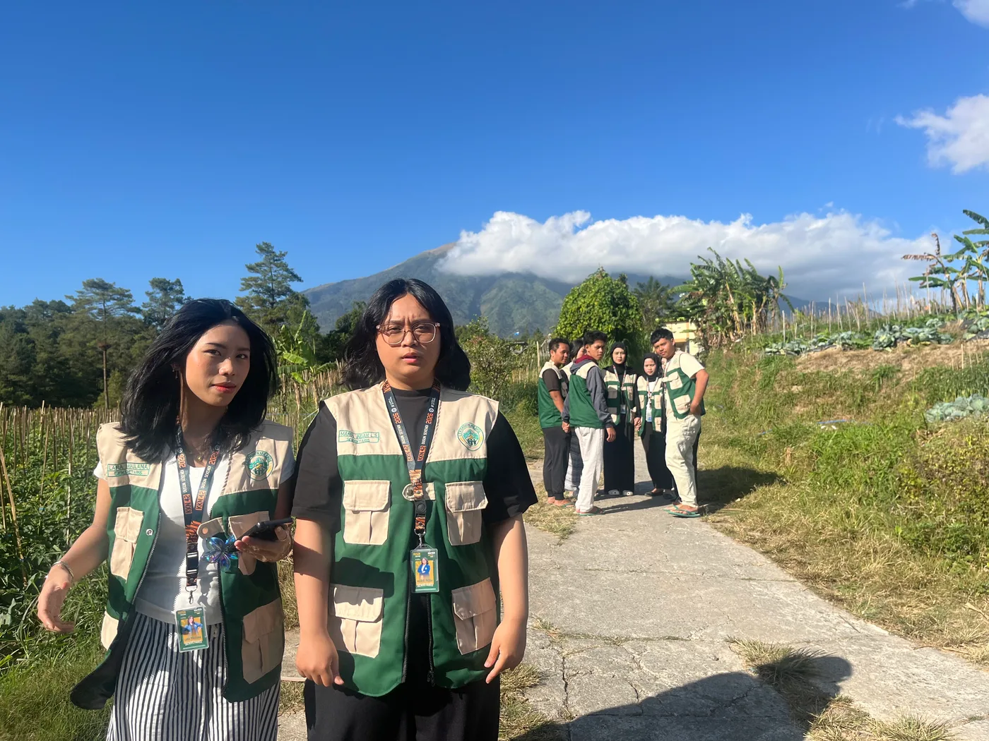Anggota KKN berjalan di jalan dusun dengan ladang dan gunung di bawah langit biru