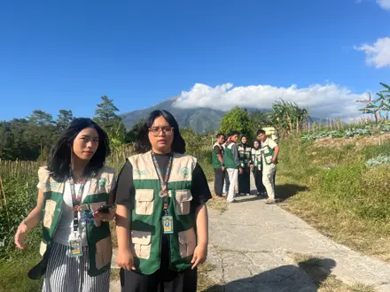 Anggota KKN berjalan di jalan dusun dengan ladang dan gunung di bawah langit biru