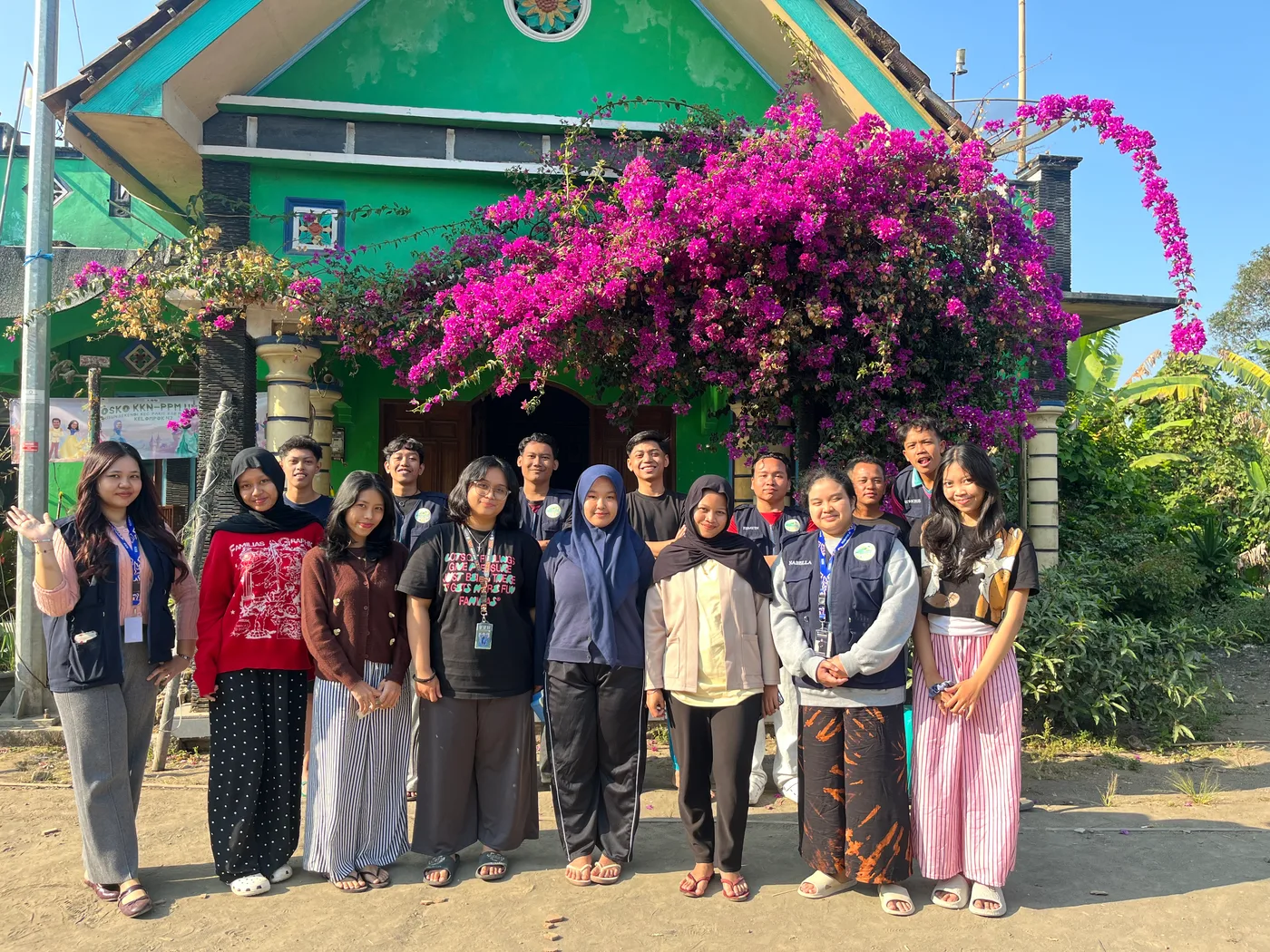Anggota kelompok berfoto bersama warga di depan posko hijau dan bunga bugenvil
