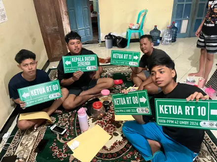 Empat mahasiswa menunjukkan papan hijau bertuliskan ketua RT di dalam posko
