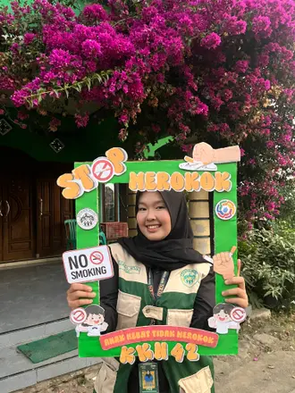 Seorang anggota KKN tersenyum memegang bingkai kampanye stop merokok di depan posko