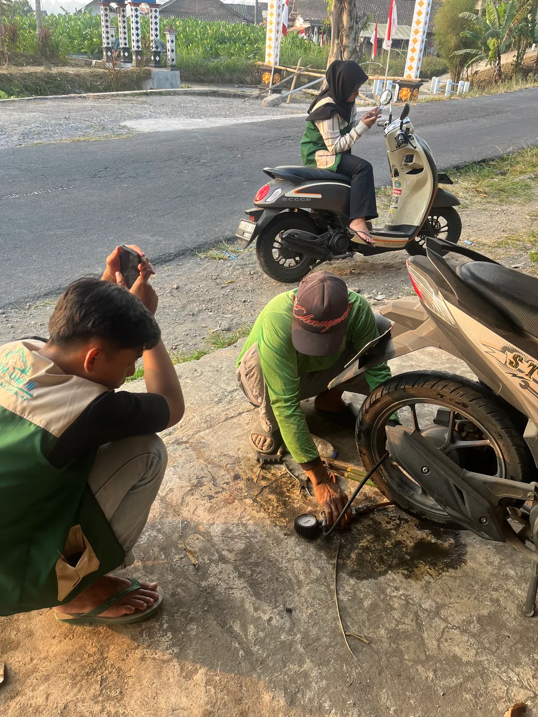 Dua mahasiswa berjongkok membantu memperbaiki ban sepeda motor di tepi jalan