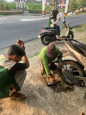 Dua mahasiswa berjongkok membantu memperbaiki ban sepeda motor di tepi jalan
