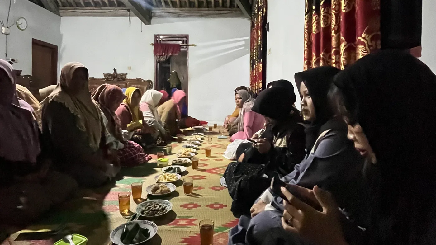 Ibu-ibu dan mahasiswa duduk memanjang di kedua sisi hidangan dalam rumah warga