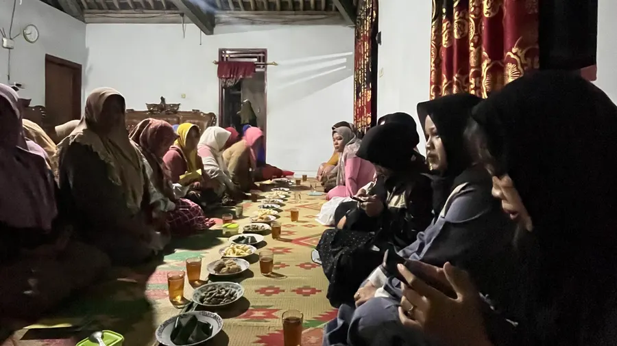 Ibu-ibu dan mahasiswa duduk memanjang di kedua sisi hidangan dalam rumah warga