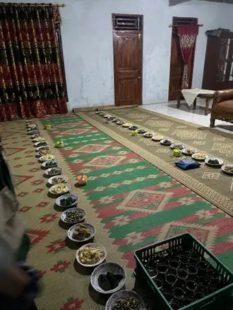 Piring-piring makanan tersusun panjang di atas tikar untuk pertemuan warga